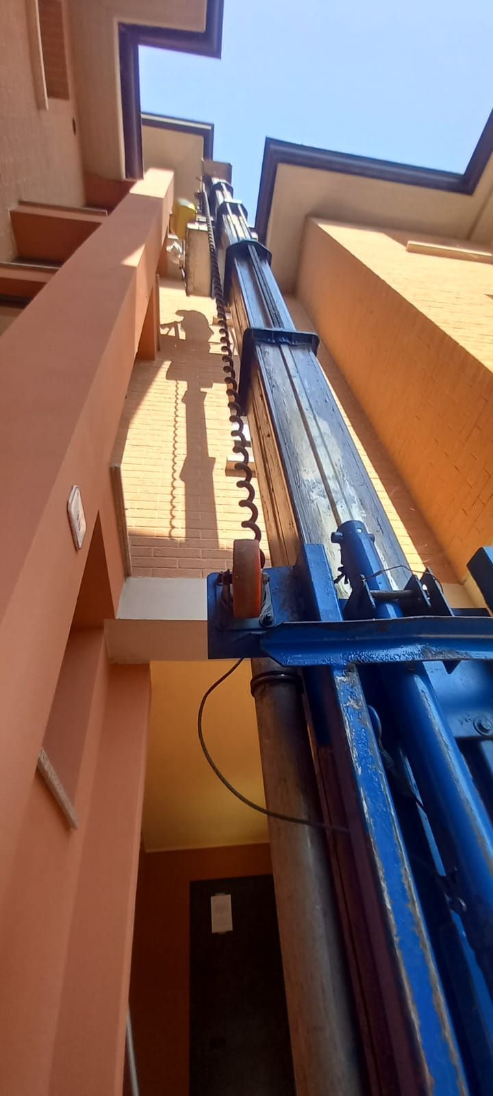 Ascensore da cantiere blu tra alti edifici arancioni, visto dal basso verso il cielo.
