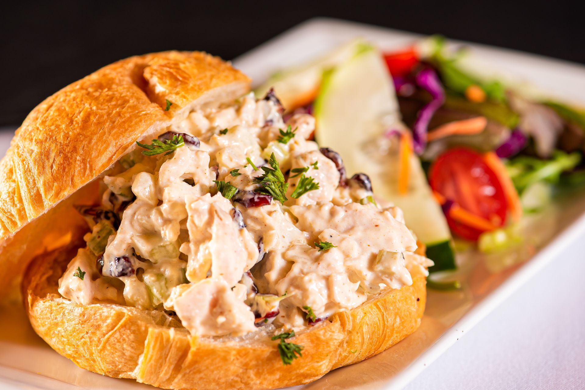 A close up of a chicken salad sandwich on a bun on a plate with a salad.
