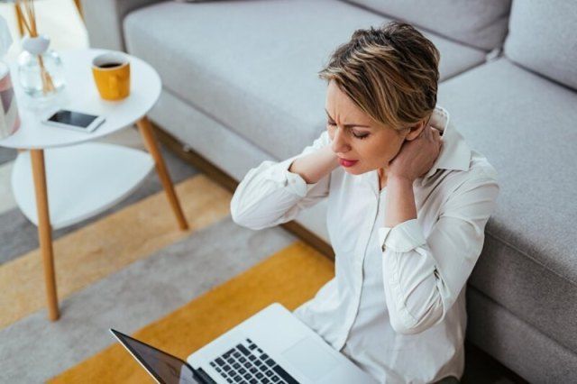 Woman holding neck in pain while working on a laptop on the floor near a couch.