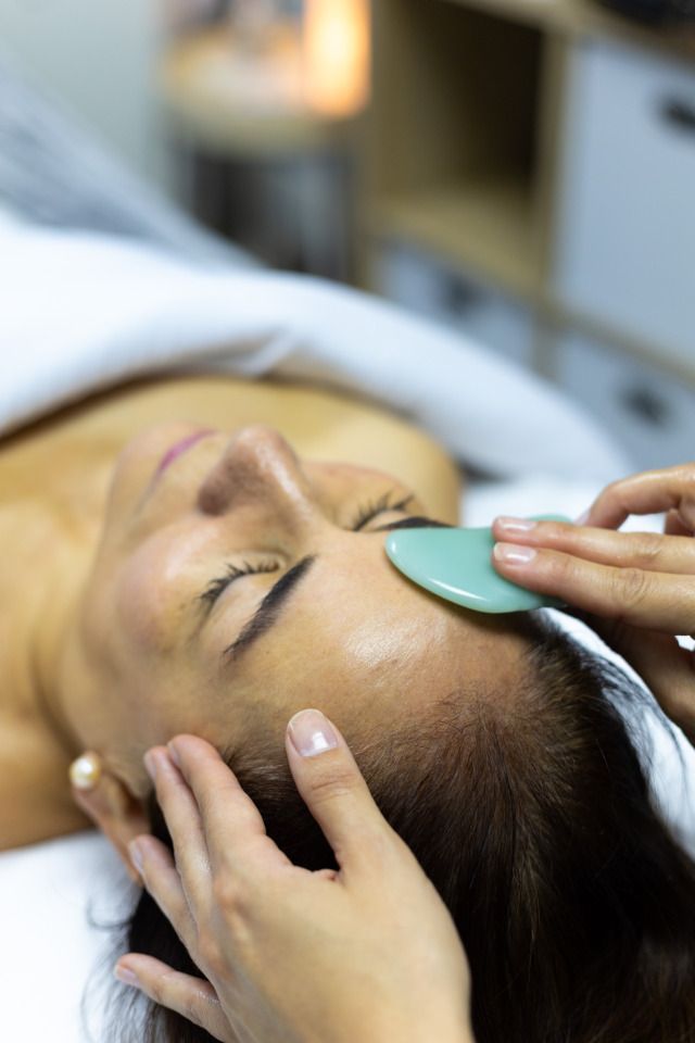 Woman receiving a facial treatment with a jade gua sha tool on her forehead in a spa setting.