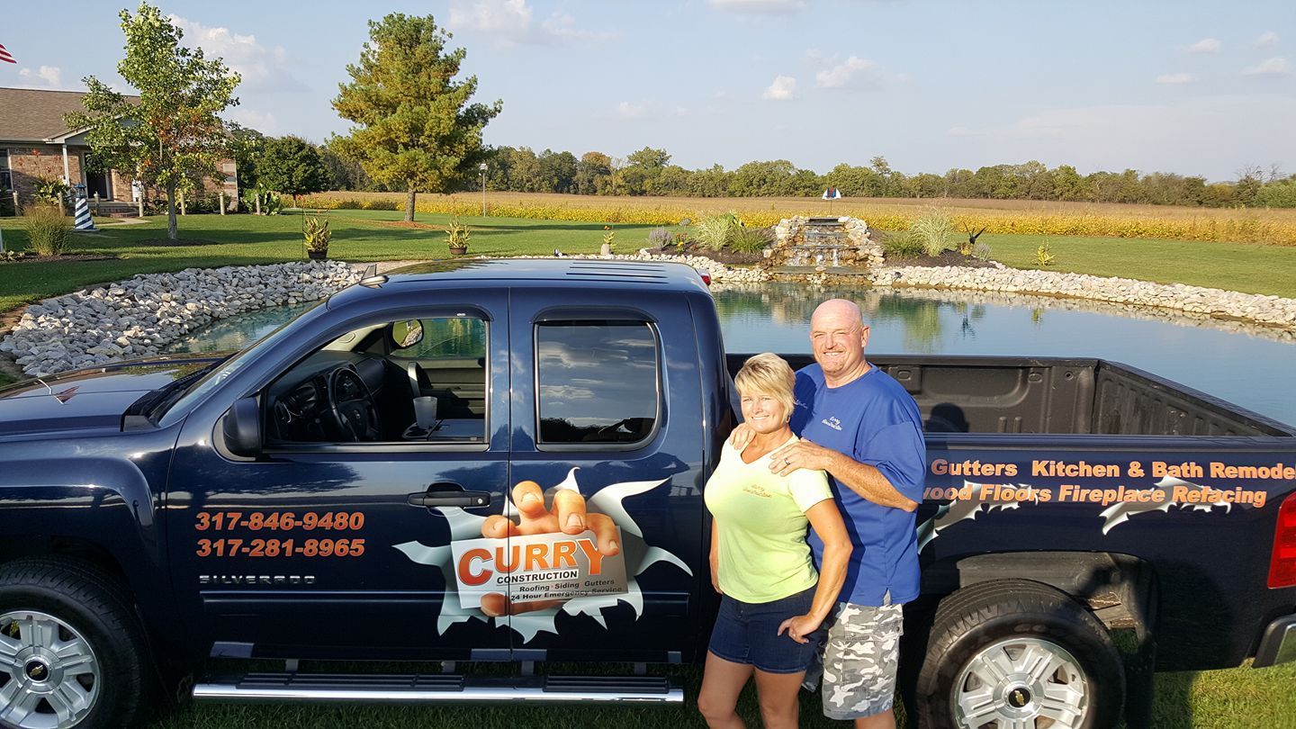 Couple poses beside a blue truck with business logo. Pond and field in background.