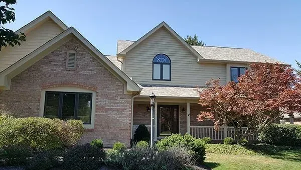 Two-story house with tan siding, brick facade, arched window, porch, and a red-leafed tree under a blue sky.