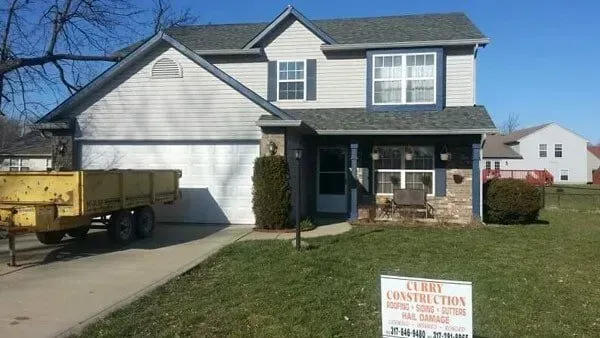 Two-story house with blue shutters and stone accents, a sign advertising construction work, and a yellow trailer.