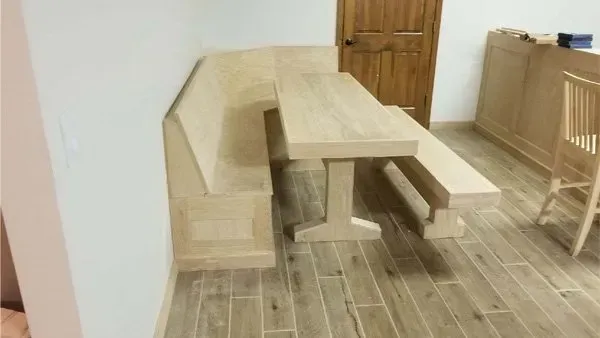 Wooden dining set: L-shaped bench, table with pedestal base, and separate bench in a room with light wood floors.