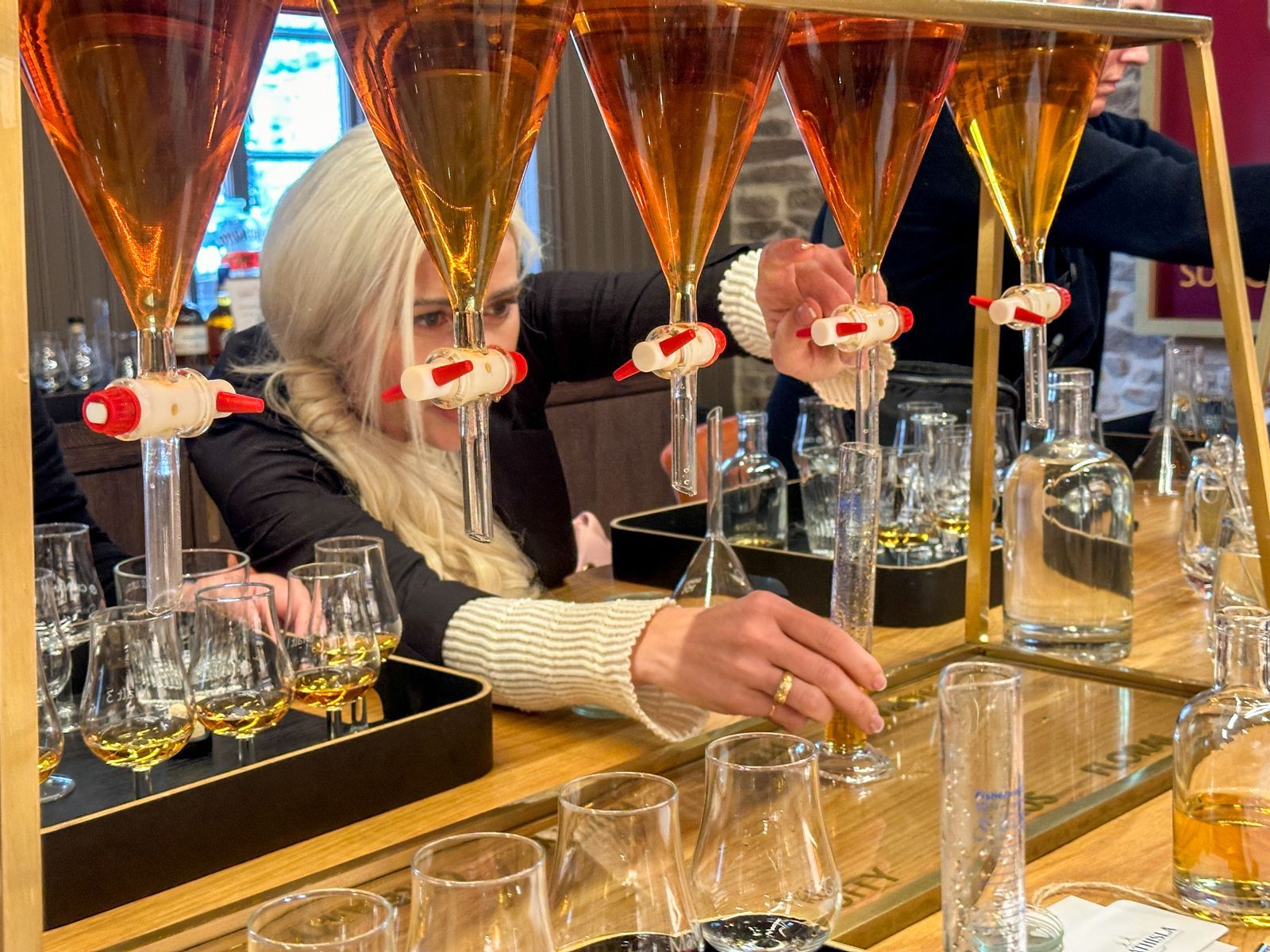 A person in a cream sweater pours golden liquid from a laboratory-style funnel into a small glass on a bar counter.