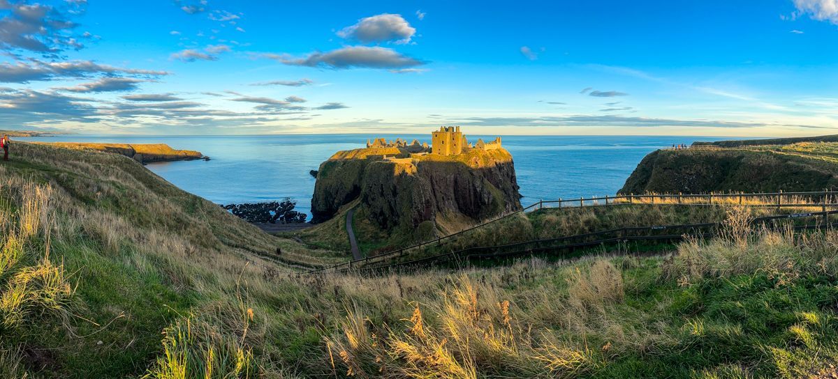 Dunnottar Castle ruins atop a dramatic cliff surrounded by the sea under a bright, partly cloudy blue sky.