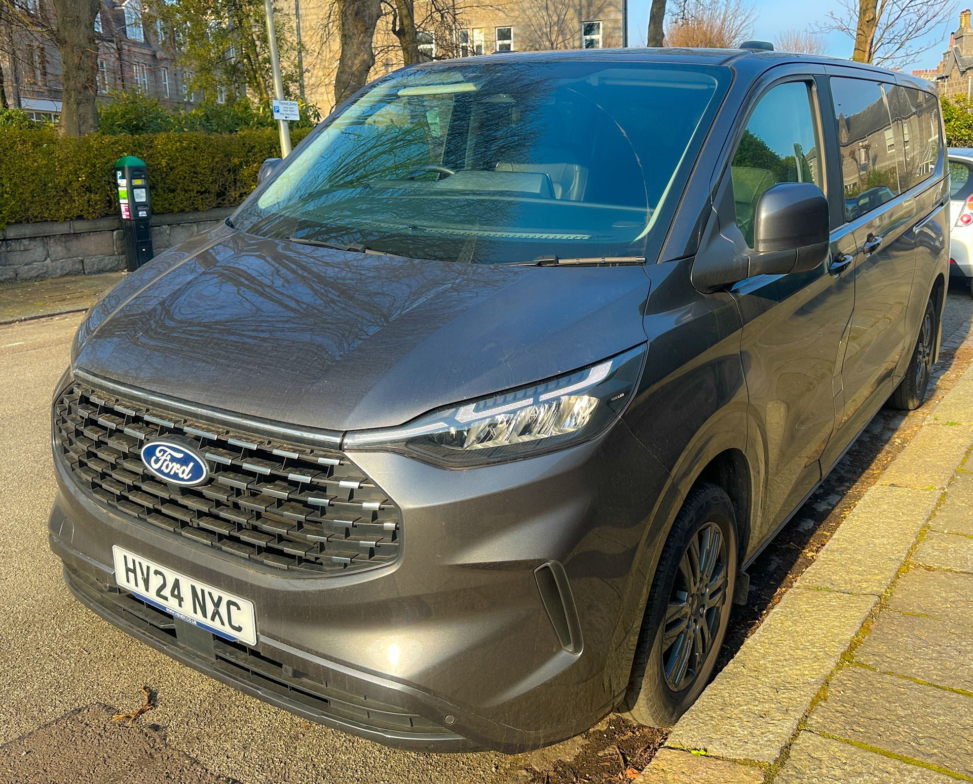 A dark grey Ford Tourneo Custom parked on a paved street beside a sidewalk with greenery in the background.