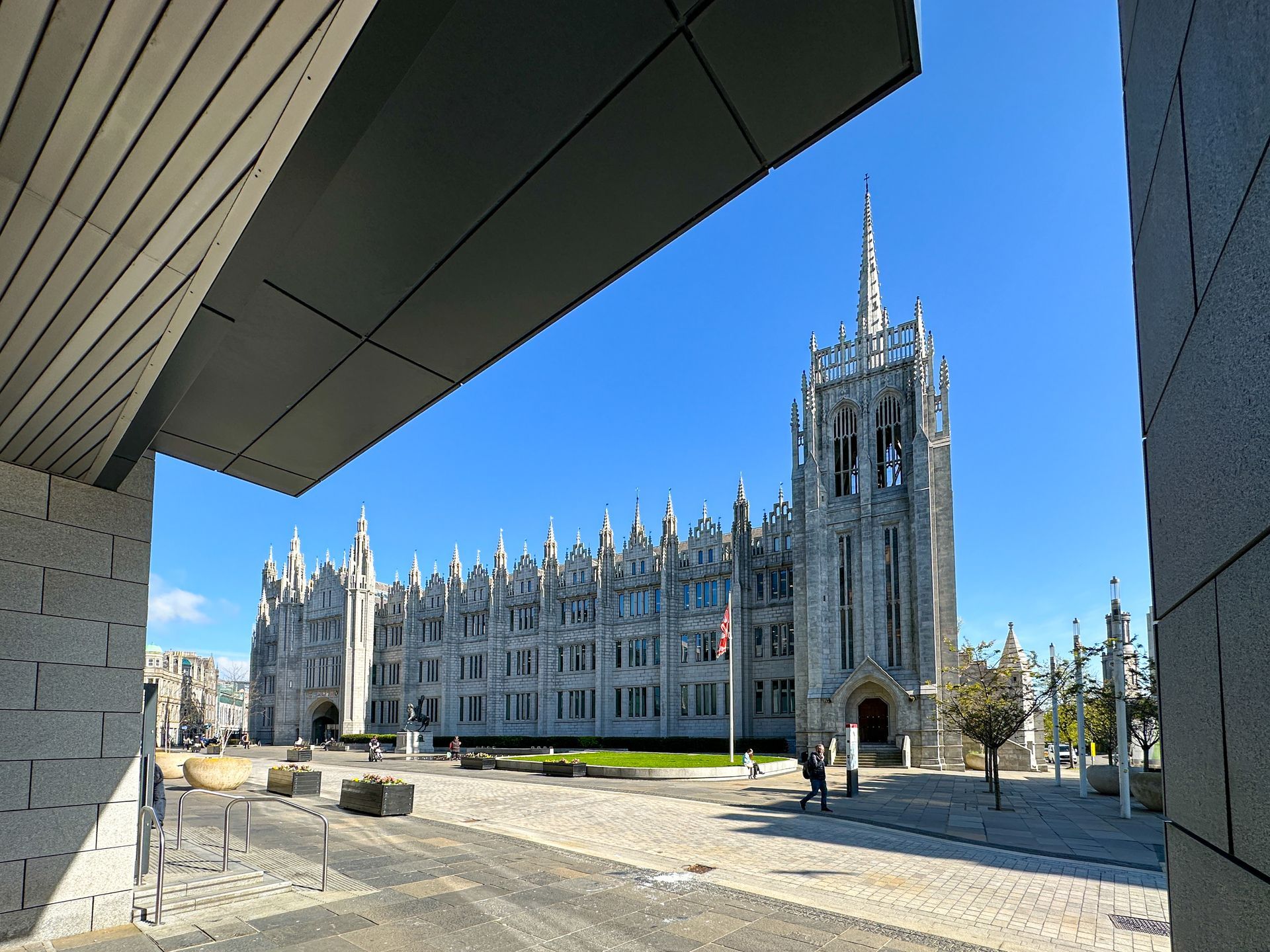 Marischal College in Aberdeen, a grand granite building with a tall spire, seen from under a modern overhang on a sunny day.
