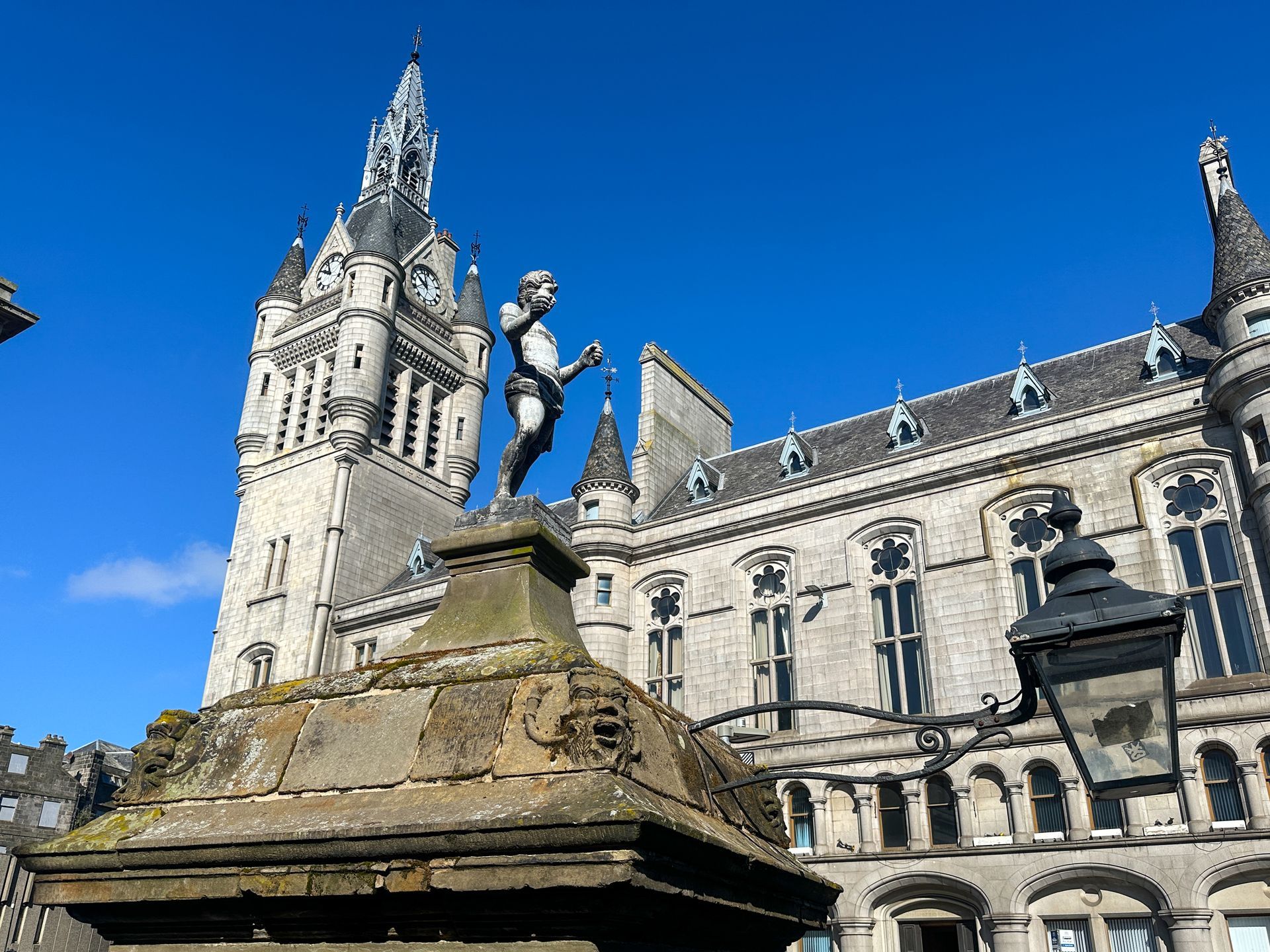 A statue of a figure stands before the ornate, light-colored stone tower and architecture of the Aberdeen Town House.