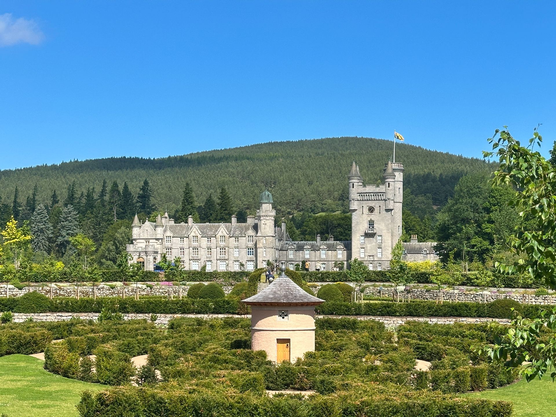Balmoral Castle with its stone tower stands against a green, forested hill, overlooking a garden with a small gazebo.