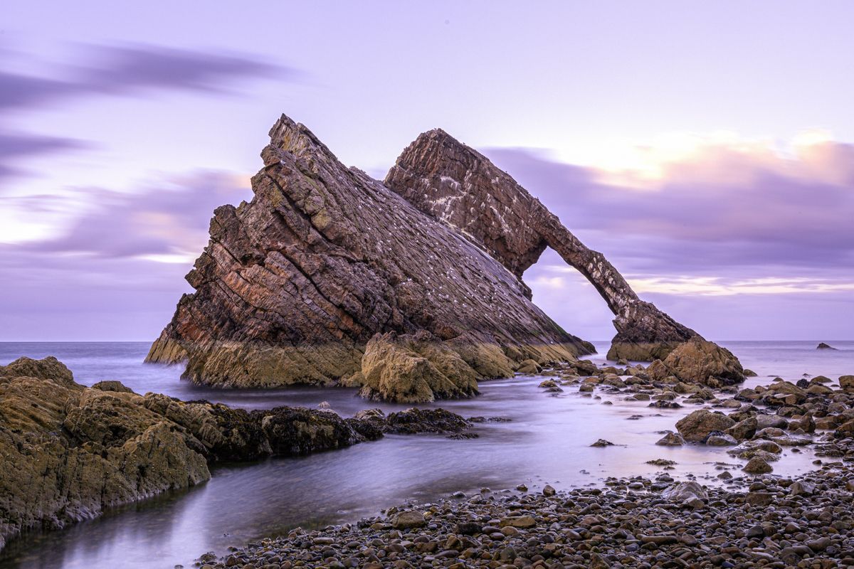 Bow Fiddle Rock in Scotland stands in calm water under a purple twilight sky, its jagged arch silhouette rising above sea.