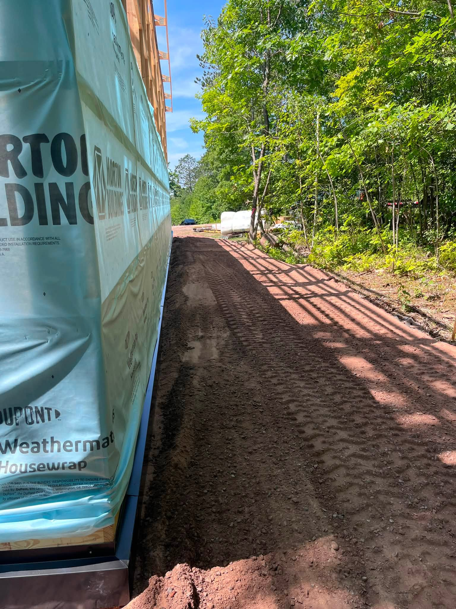 Narrow dirt path beside a blue construction tarp wall, with trees and sunlight ahead