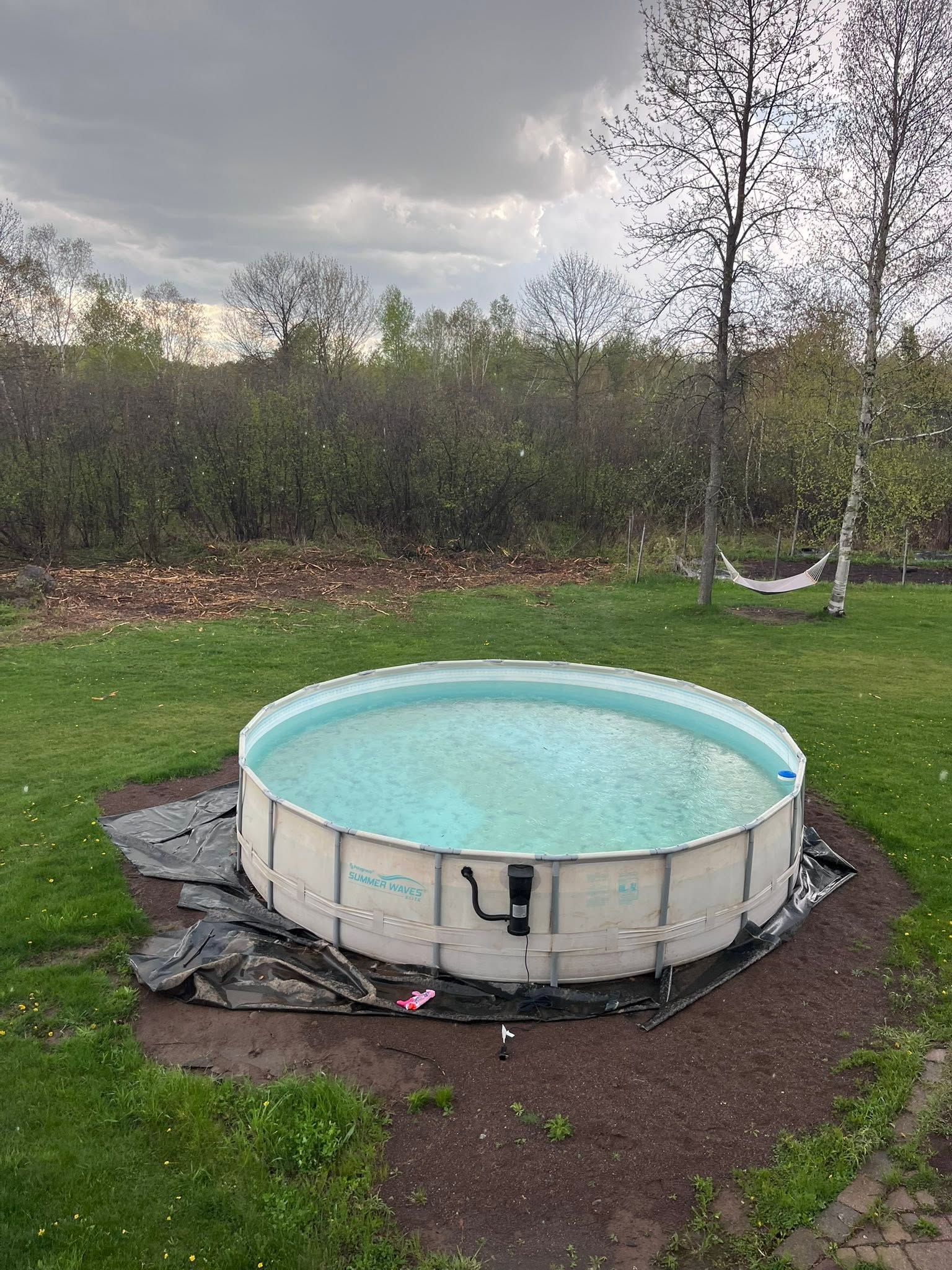 Small above-ground pool in a backyard with cloudy sky and a hammock between trees.