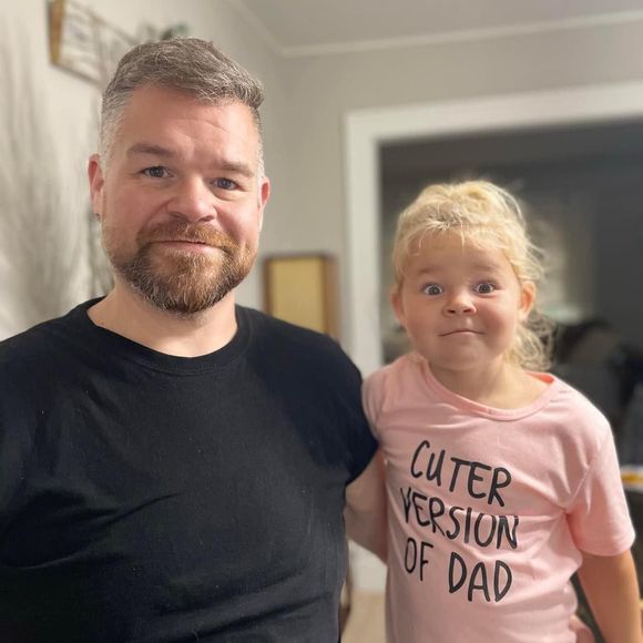 Man and child posing indoors; child wears pink shirt reading “CUTER VERSION OF DAD.”