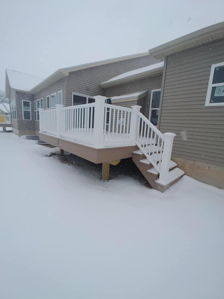 Snow-covered white deck with railing and steps attached to a tan-sided house during snowfall.