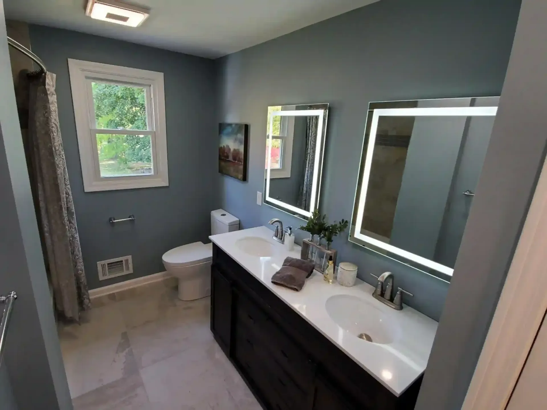 Bathroom with double vanity, illuminated mirrors, toilet, and window with outdoor view.