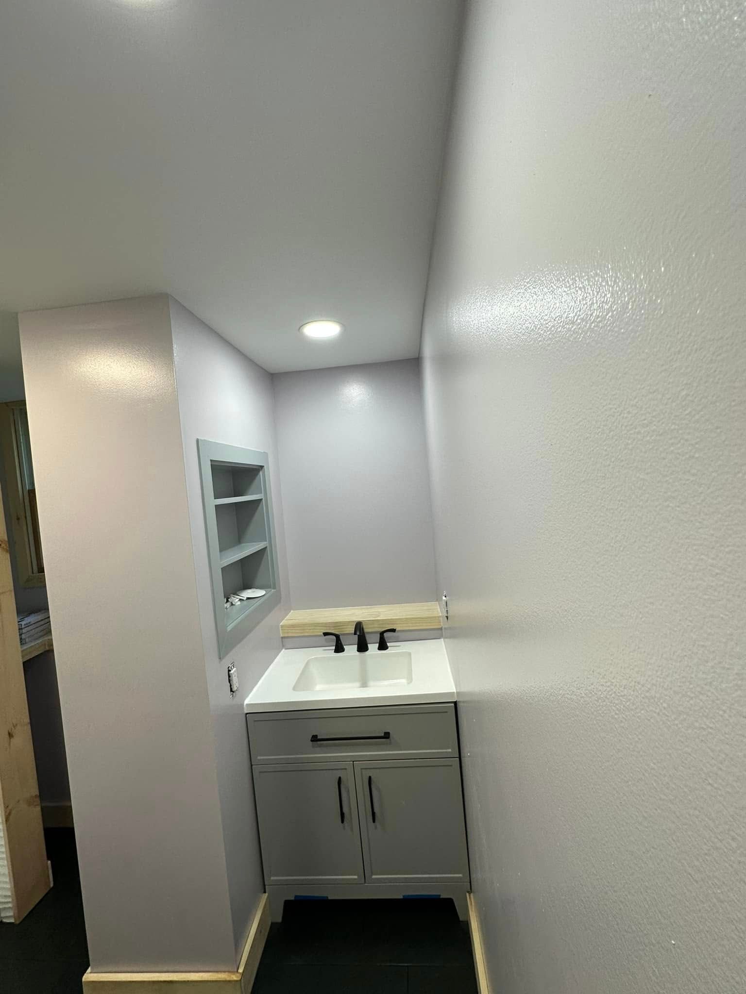 Small, light purple bathroom with white vanity, built-in shelving, and black sink.