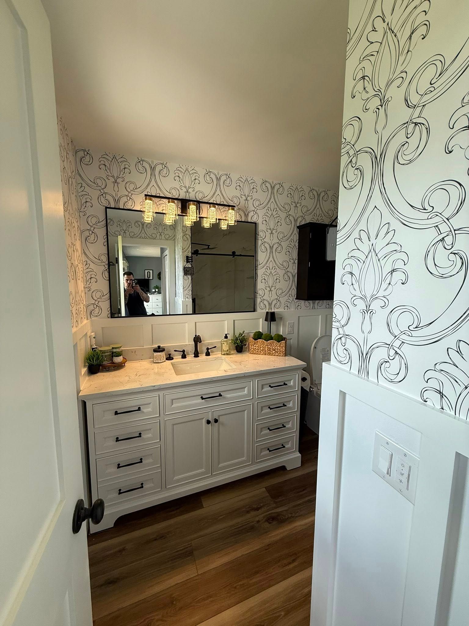 Bathroom with white vanity, patterned wallpaper, large mirror, and wood-look flooring.