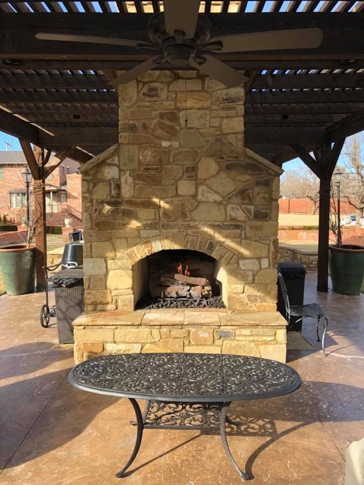 Stone outdoor fireplace with fire, under a pergola, table in foreground. Stone outdoor fireplace with fire, under a pergola, table in foreground.