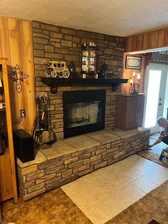 Stone fireplace with black interior and wooden mantel, decorated with figurines. Fireplace hearth extends out, carpet in front.