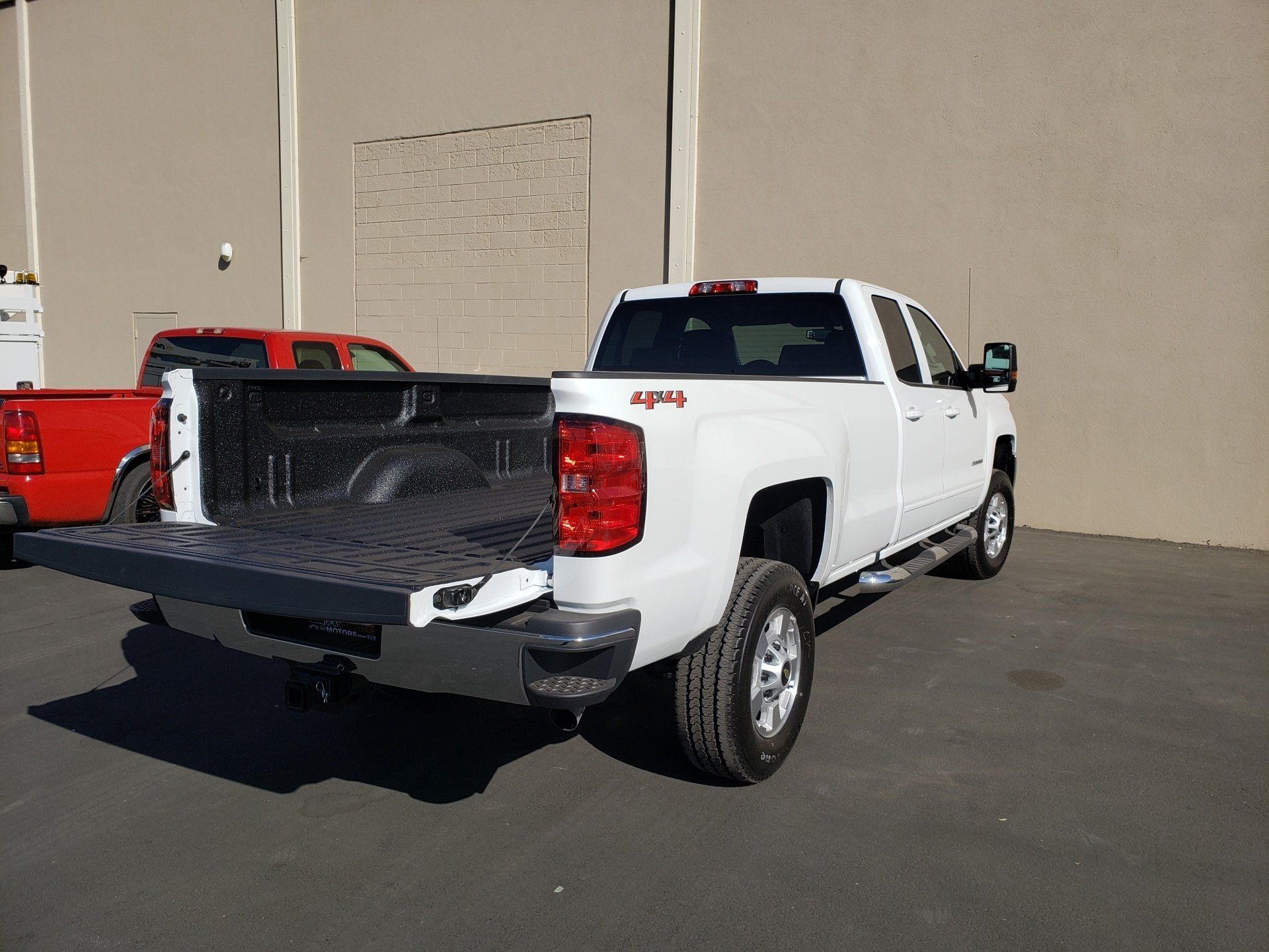 White pickup truck with open tailgate parked beside a red truck outside a beige building