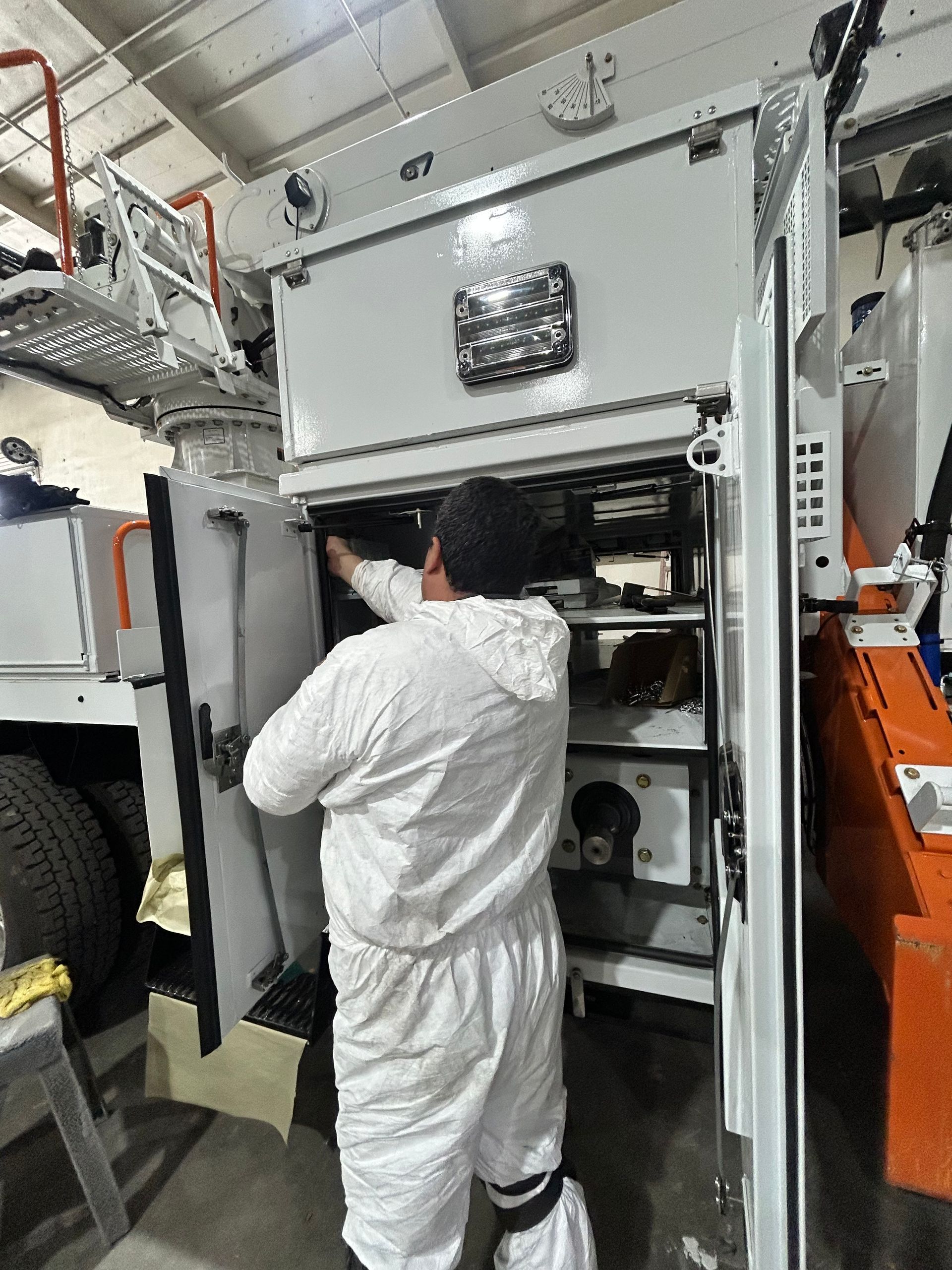 Worker in white coveralls inspecting equipment inside a control cabinet on a ship or industrial vessel.