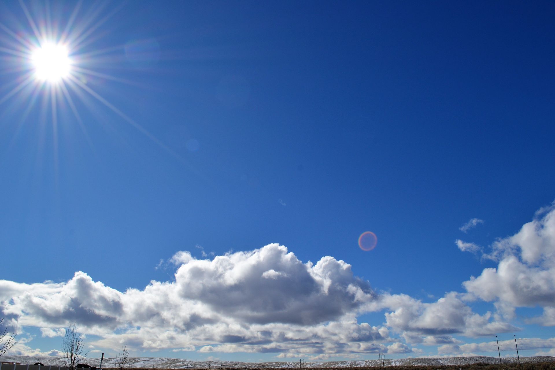 Bright blue sky with the sun, scattered clouds, and a faint rainbow over a distant horizon