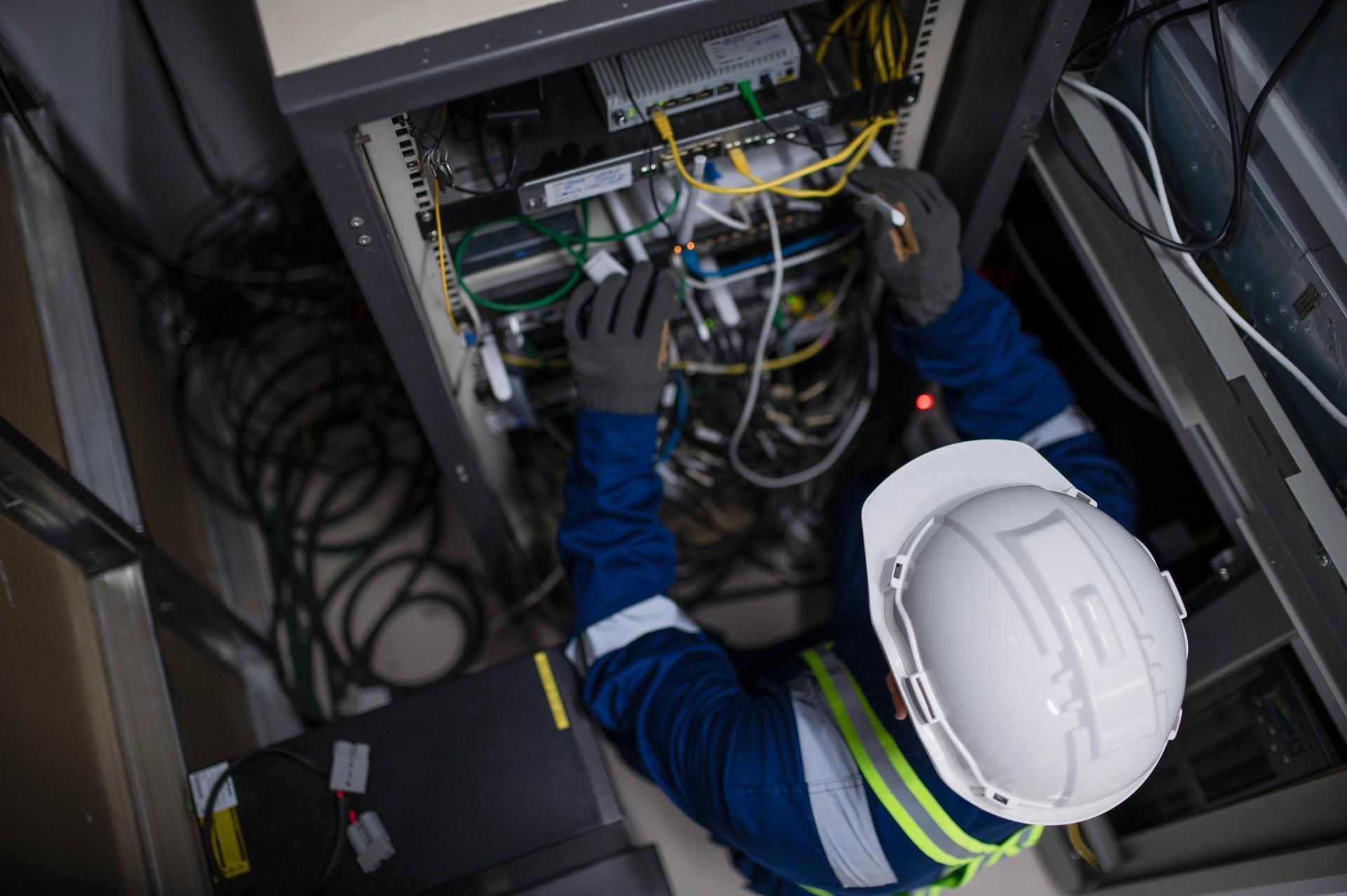 Technician in blue work suit and hard hat, working on server cables inside a rack.