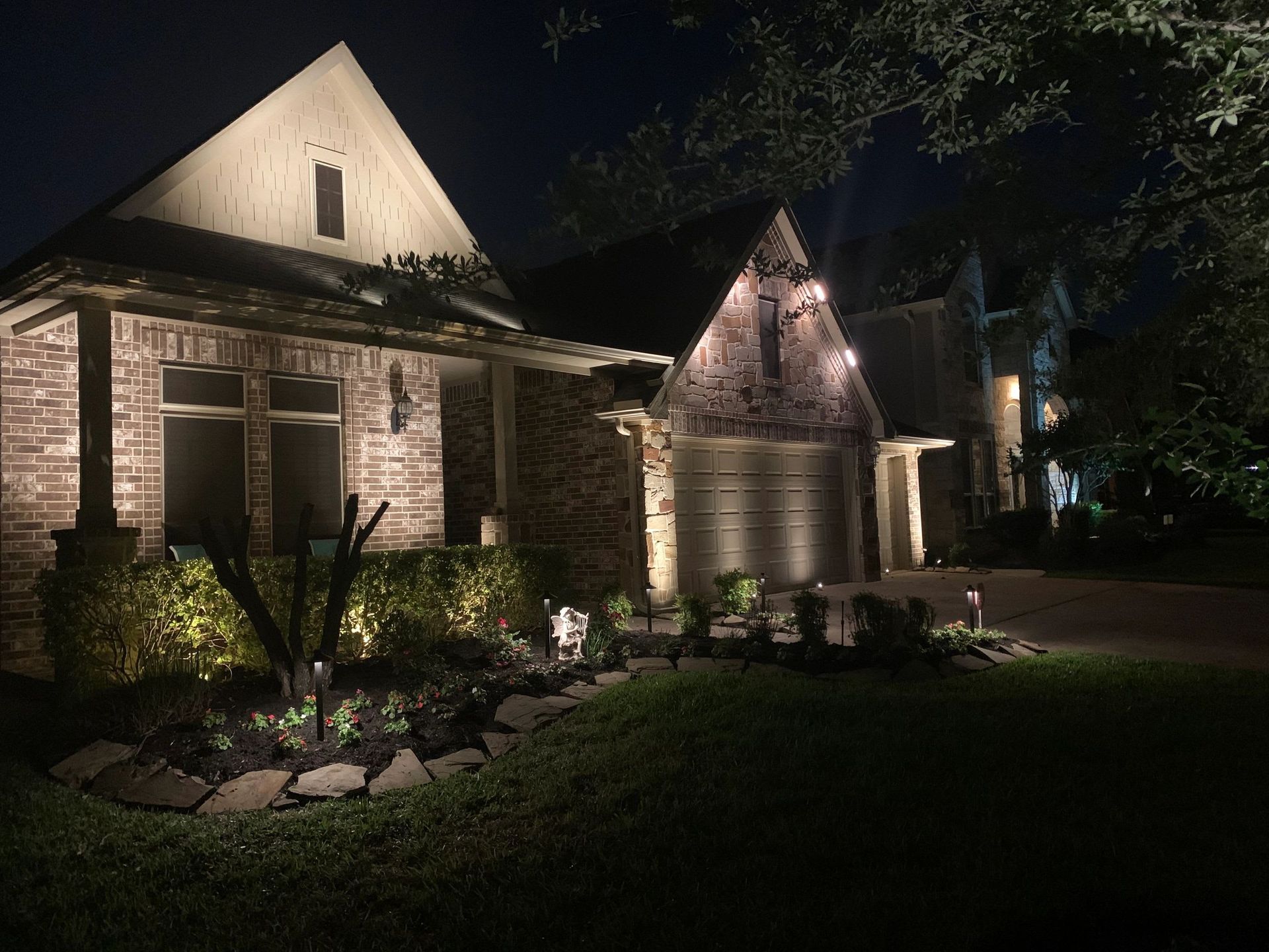 House exterior illuminated by landscape lighting at night, showing brick facade, garage, and landscaping.