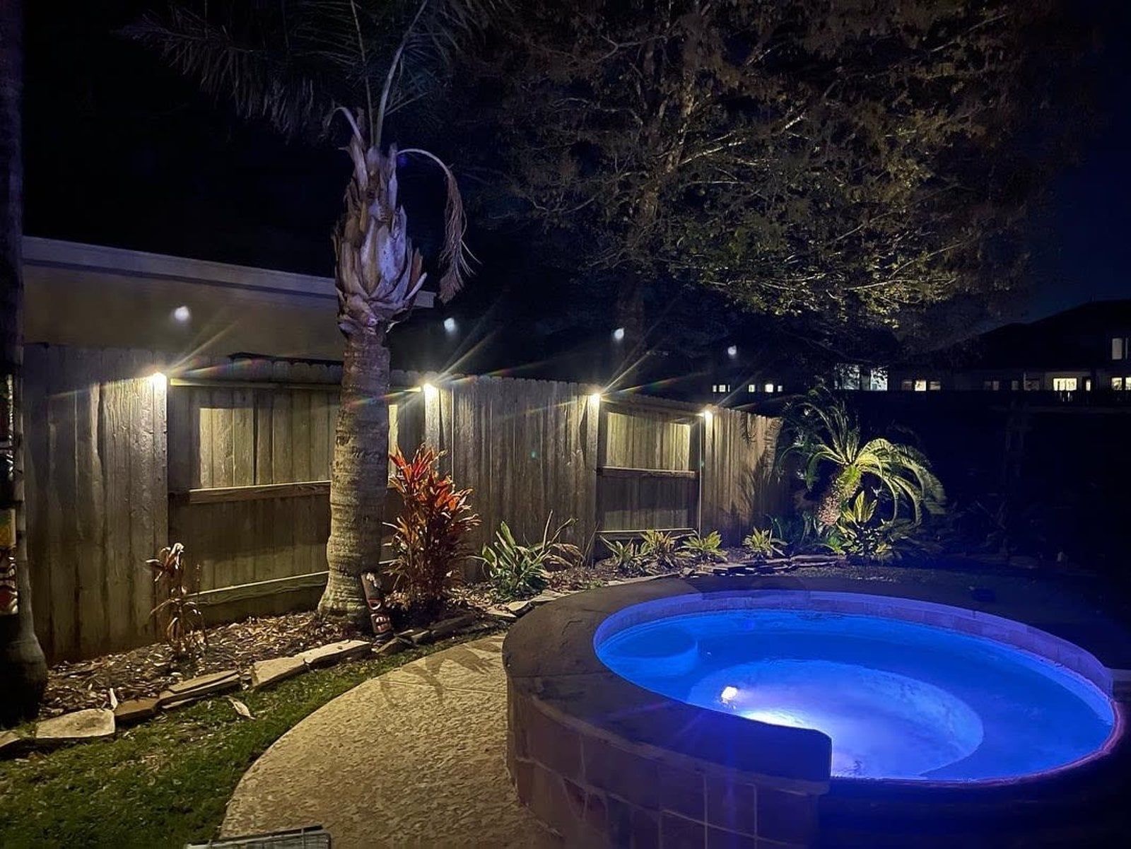 A lit-up hot tub at night with a wooden fence and landscaping.