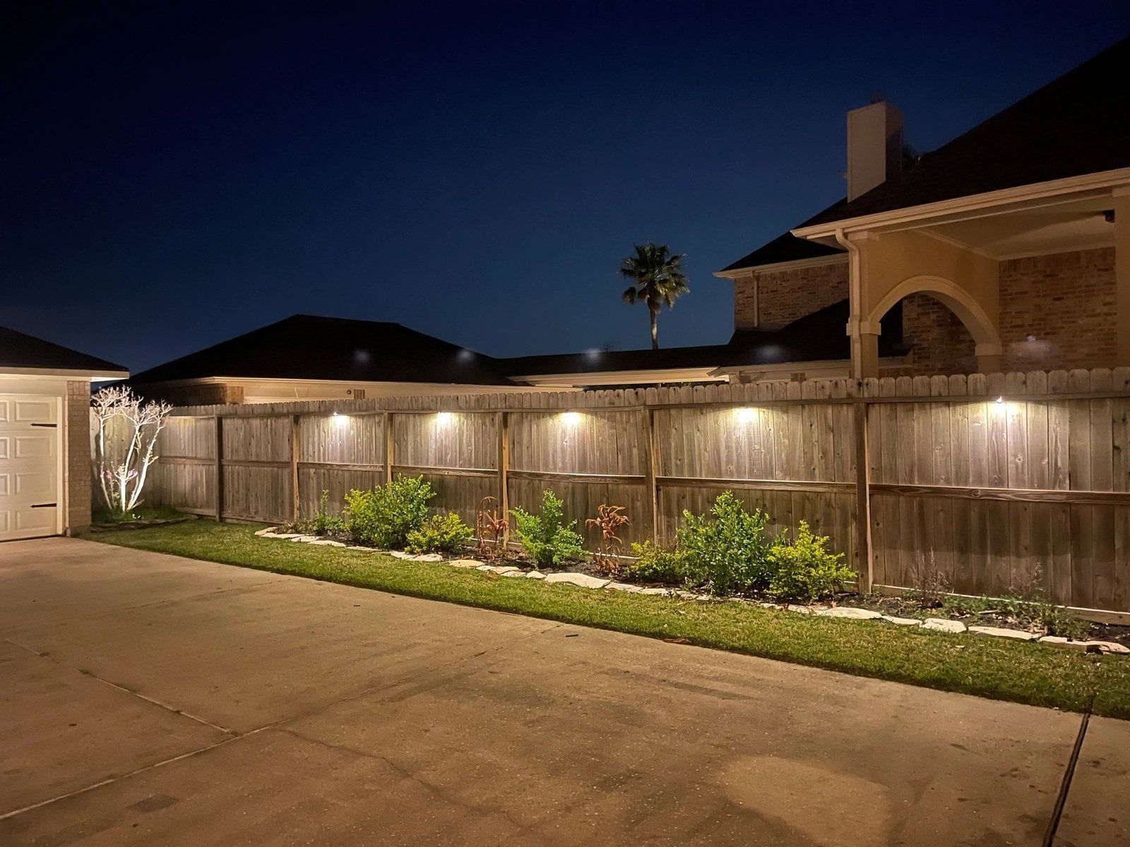 Wooden fence with lights, bushes, and a driveway at night.