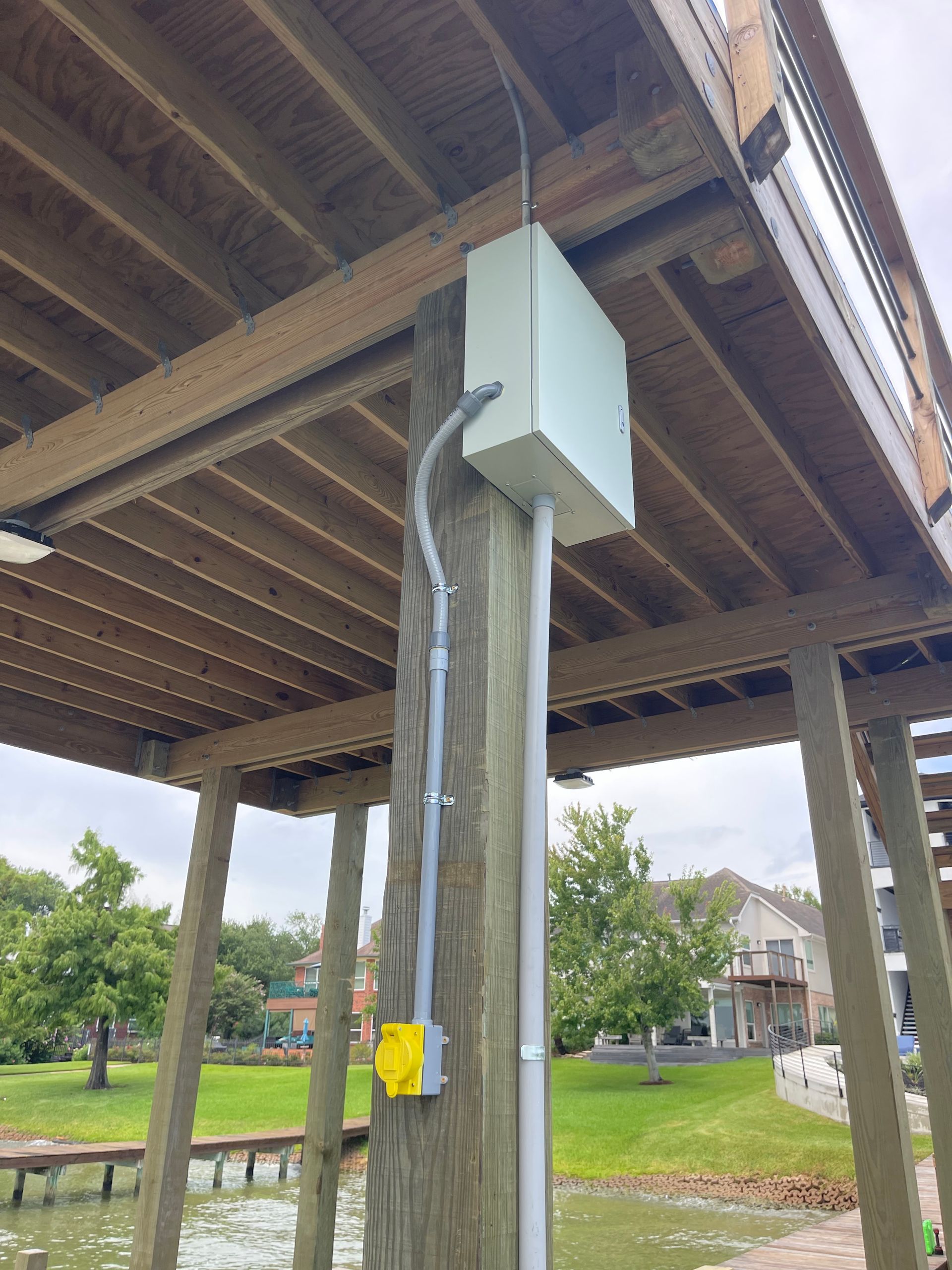 Electrical box and conduit on a wooden post under a dock. Outlet box on the post, green grass, water, and homes in the background.