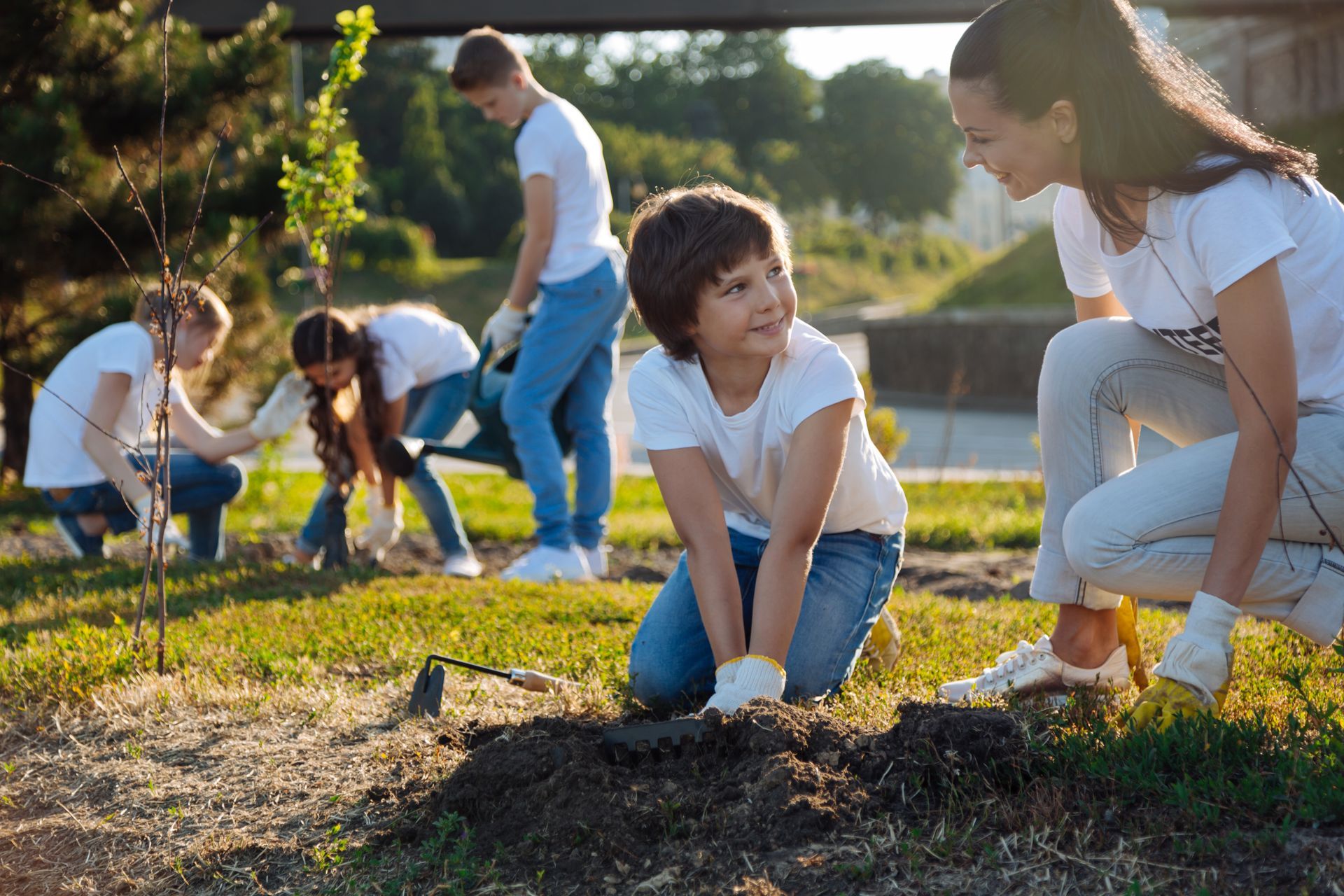 Children planting saplings in a sunny park, working together with gloves and small shovels.