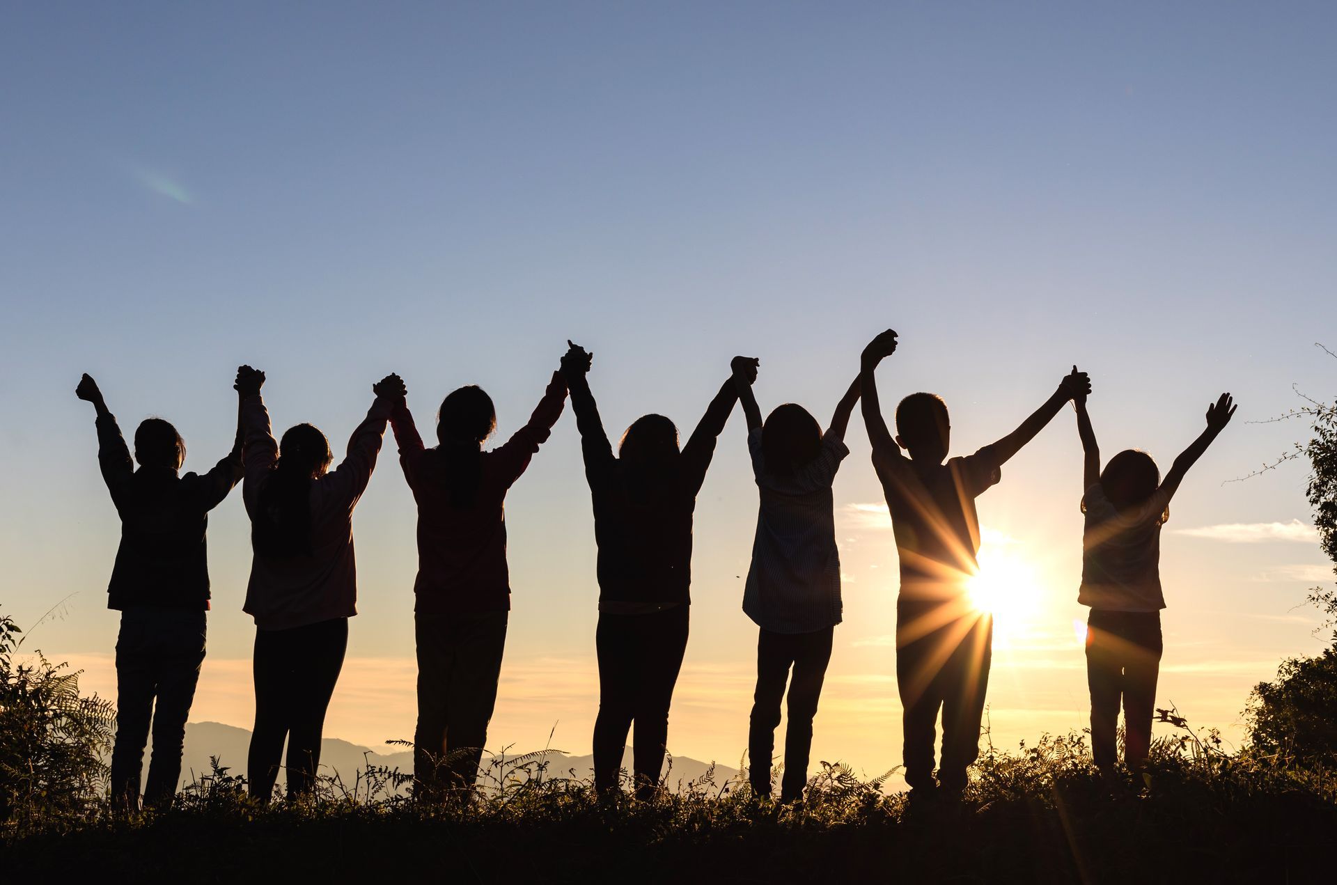 Silhouetted people holding hands with raised arms at sunset on a grassy hill