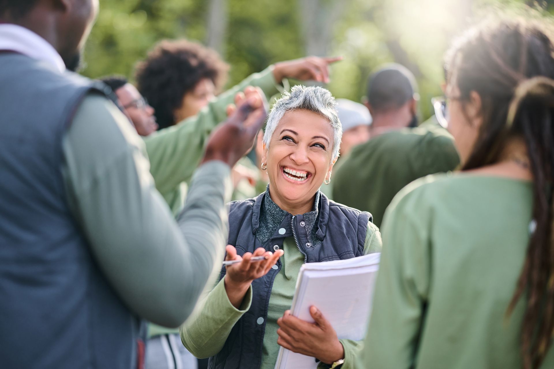 Smiling person speaking outdoors with a small group, holding papers and gesturing during a discussion