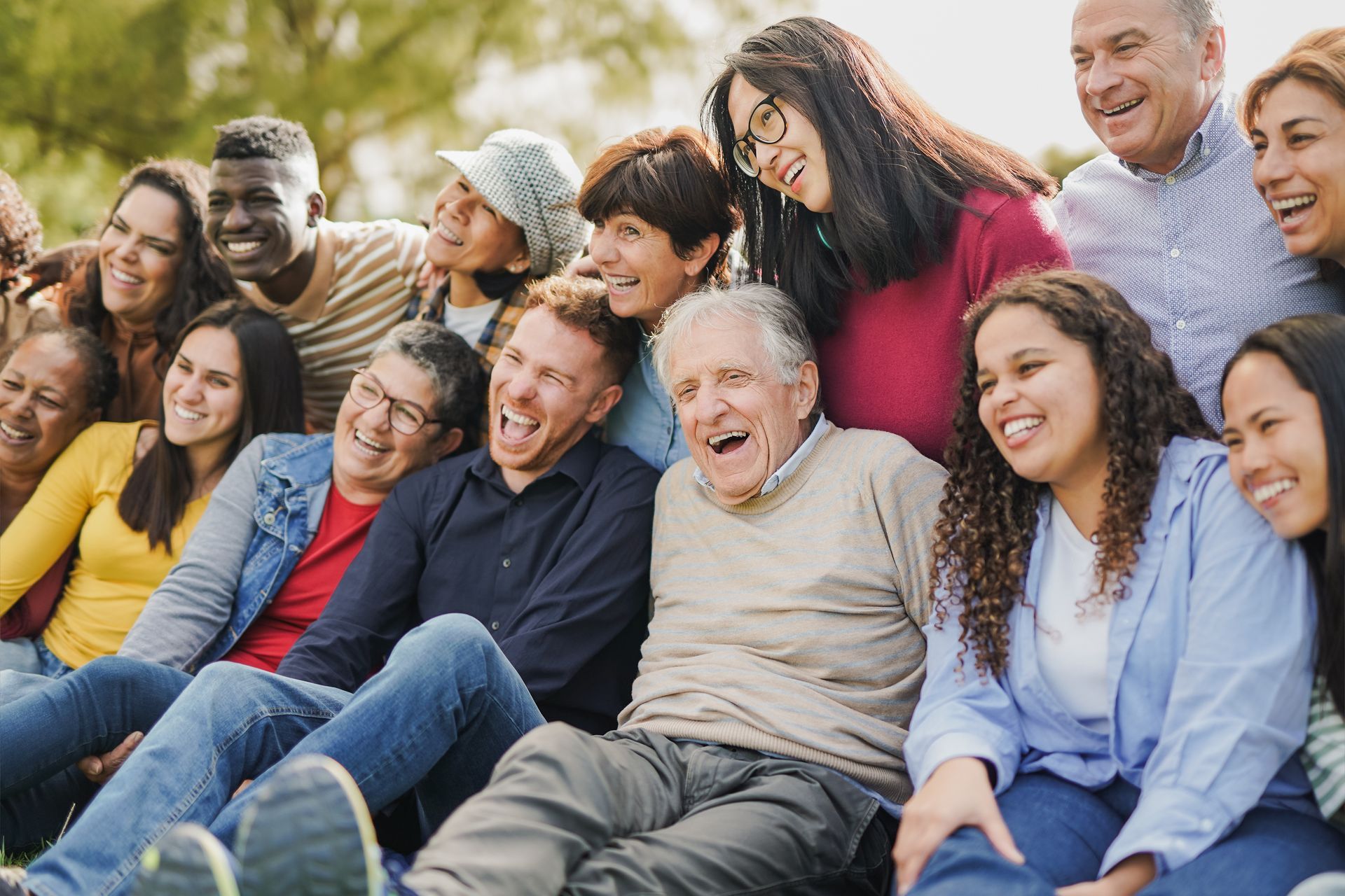 A group of friends sitting outdoors on grass, smiling and laughing together in a sunny park.
