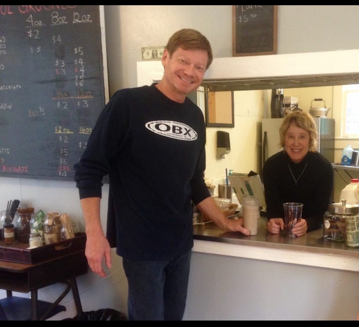Man and woman at a cafe counter. Man smiles and leans forward. Woman smiles from behind counter.