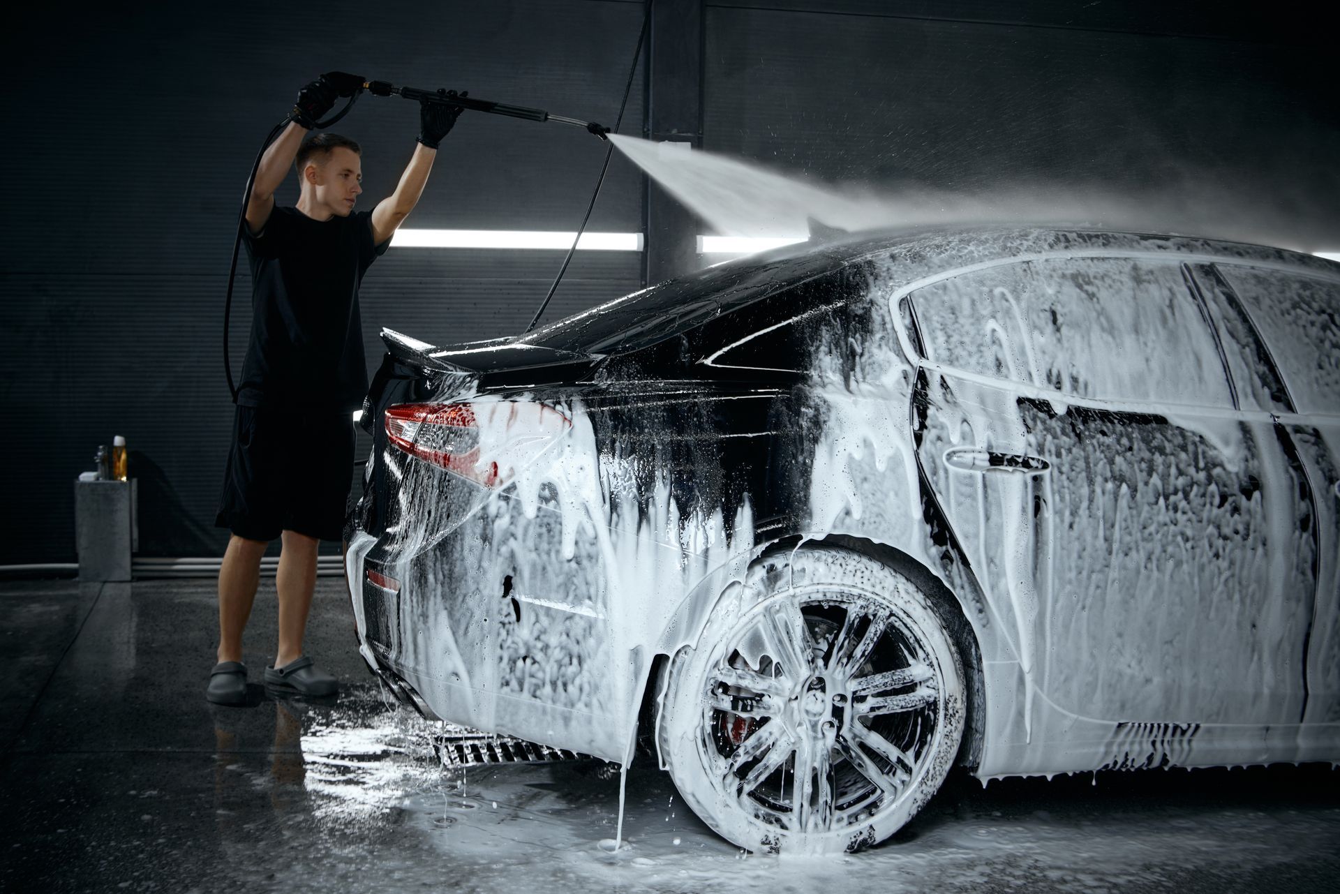 Person washing a black car with foam in a dim auto detailing bay