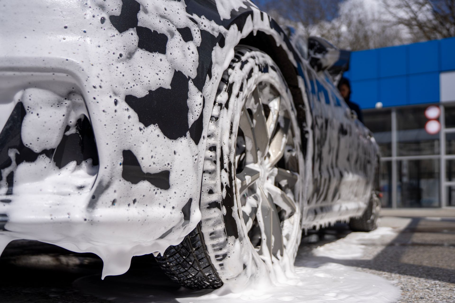 Car covered in soap foam being washed beside a building, with a wheel in close-up