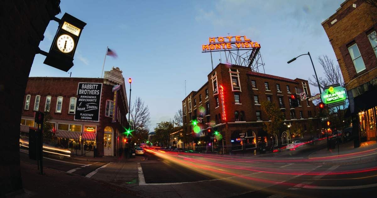 Downtown street at dusk with lit buildings, clock tower, and blurred car lights streaking through the intersection