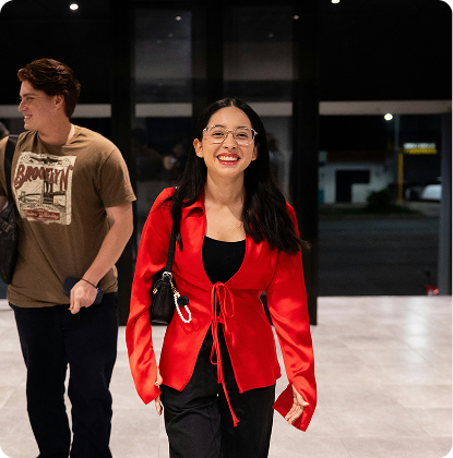 Woman in red blazer and black pants walking indoors, smiling, with a man standing behind her.