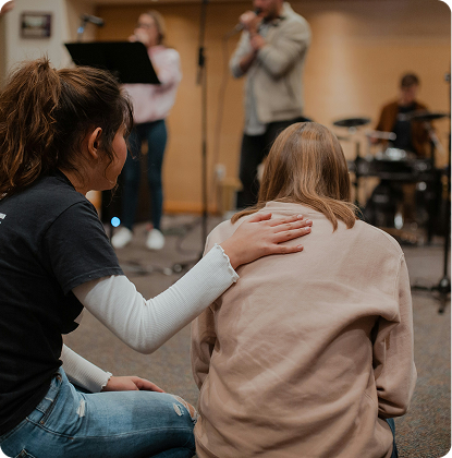 Two people sit in a church service, one comforting the other, while musicians perform on stage.
