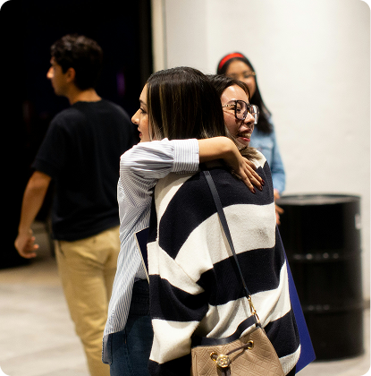 Two people hugging in a gallery, with others standing in the background.
