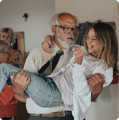 Elderly man in glasses holding a smiling child indoors during a playful dance.