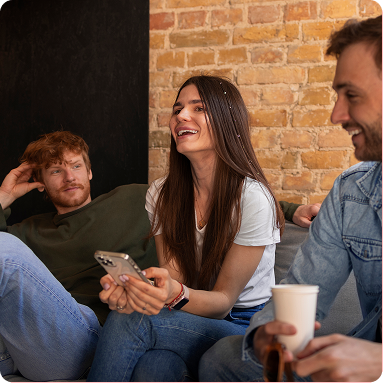Friends chatting and laughing on a couch in a brick-walled room, one holding a phone and another a coffee cup