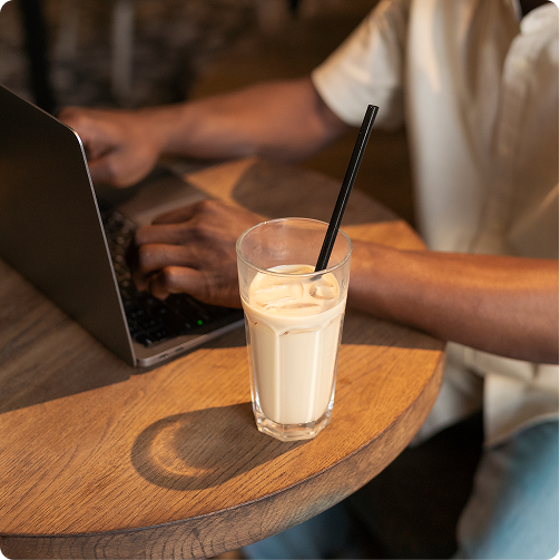 Laptop and iced drink on a wooden table, with a person typing in the background.