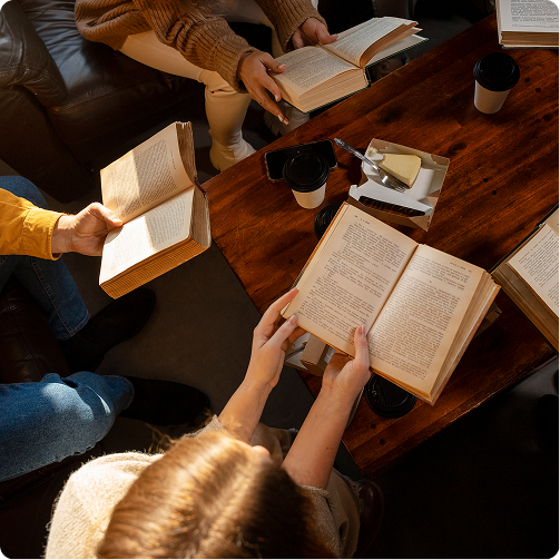 People seated around a wooden table, reading open books and discussing in warm sunlight