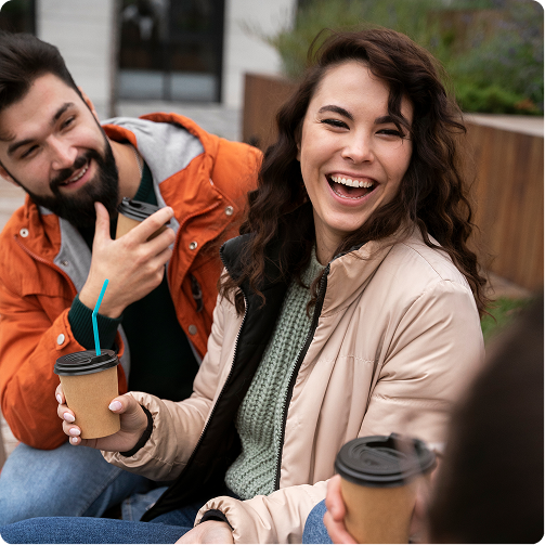 Two friends smiling outdoors on a bench, holding coffee cups, one in an orange jacket