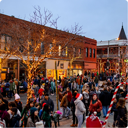 Crowded outdoor holiday market at dusk with festive lights, brick buildings, and a decorated Christmas tree