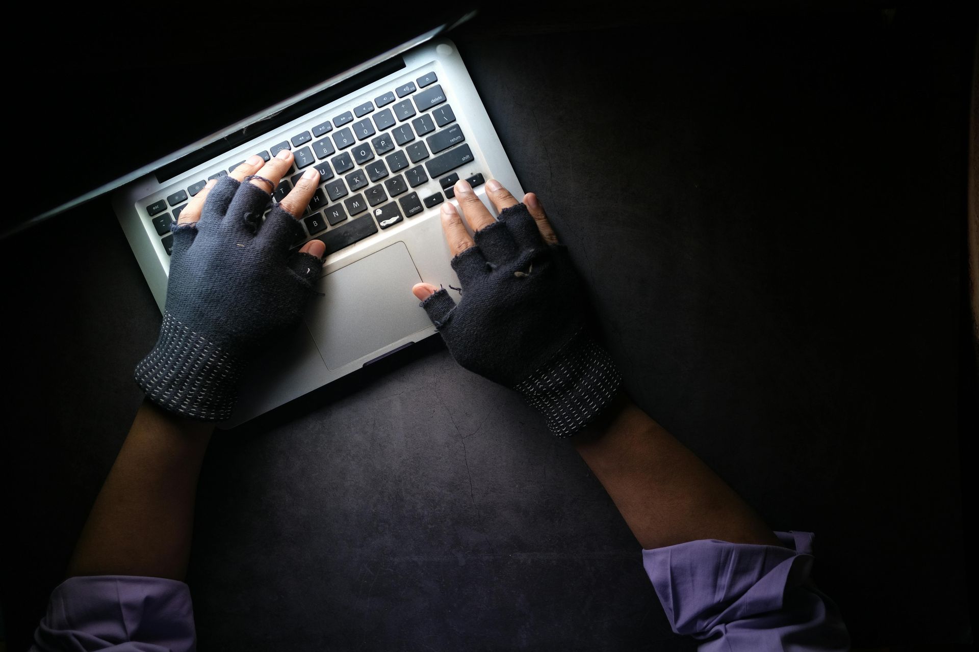 Hands wearing fingerless gloves typing on a laptop in a dimly lit setting.